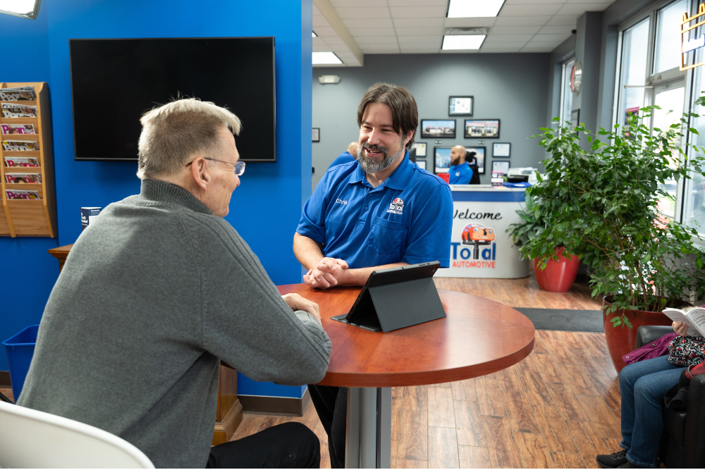 customer questions, auto repair in Buffalo, NY at Total Automotive. Service advisor at an auto repair shop speaking with a customer across a round table with a tablet, inside a modern, welcoming waiting area.
